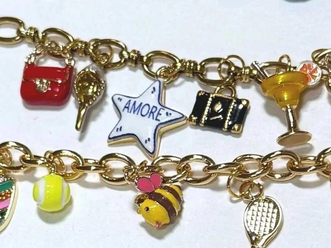 Gold chain bracelet with various charms including a star with 'AMORE' written on it, a red purse, a yellow cocktail glass, a black gift box, a pink and yellow bee, a tennis ball, a pink heart, and a gold tennis racket.