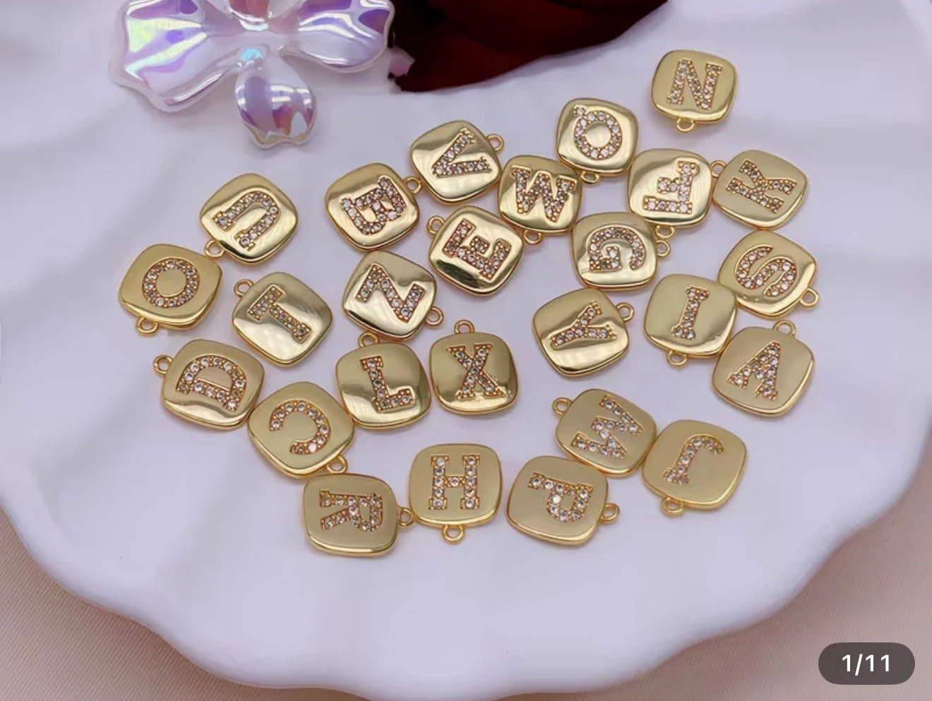 Gold-colored jewelry charms with individual letters of the alphabet, decorated with small rhinestones, arranged on a white fabric surface.