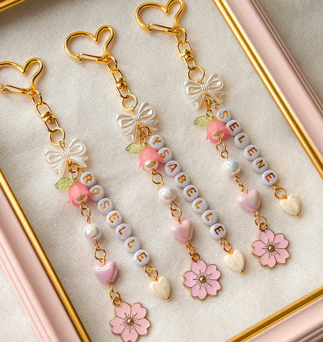 Three pairs of decorative keychains with pink flowers, white beads, pink hearts, bows, and the name 'VIVIANELLE' spelled out with letter beads, displayed in a pink tray.