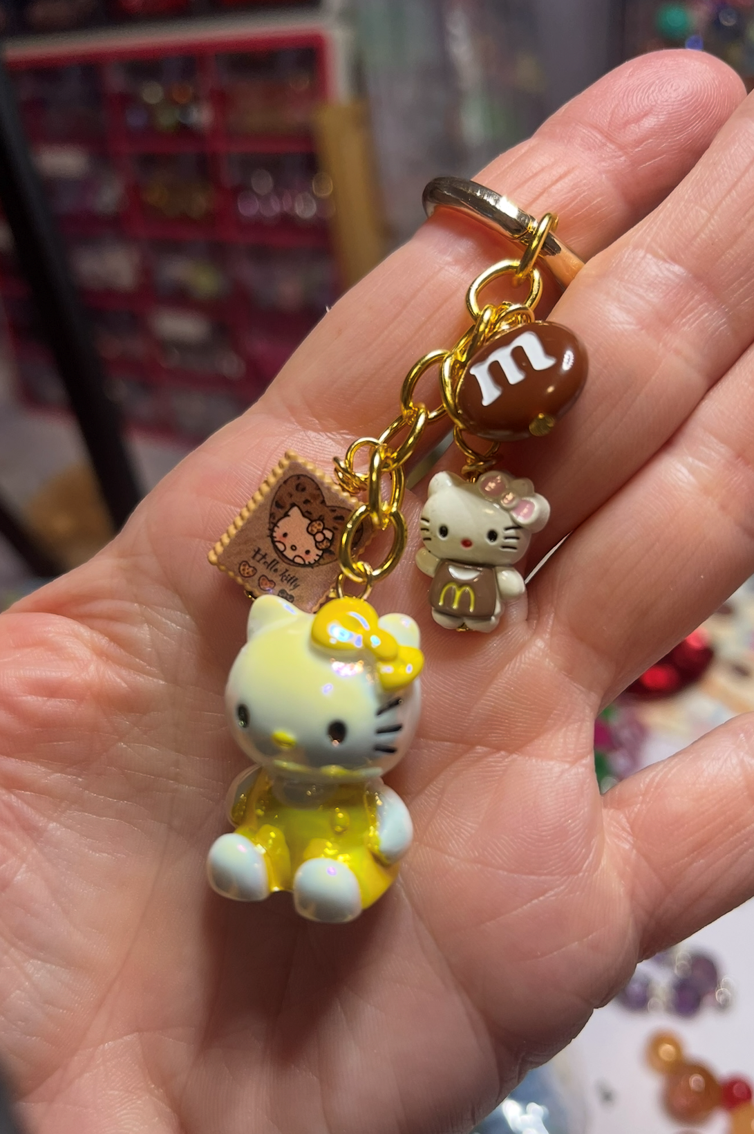 Hand holding Hello Kitty and McDonald's themed keychains, including a yellow Hello Kitty figure, a small Hello Kitty charm, a brown McDonald's M&M charm, and a Hello Kitty dressed as a McDonald's worker.