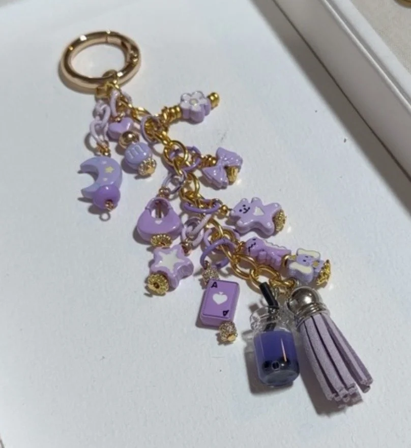 A lavender and gold charm keychain with various charms including stars and moons, a playing card, a small bottle, and a tassel.