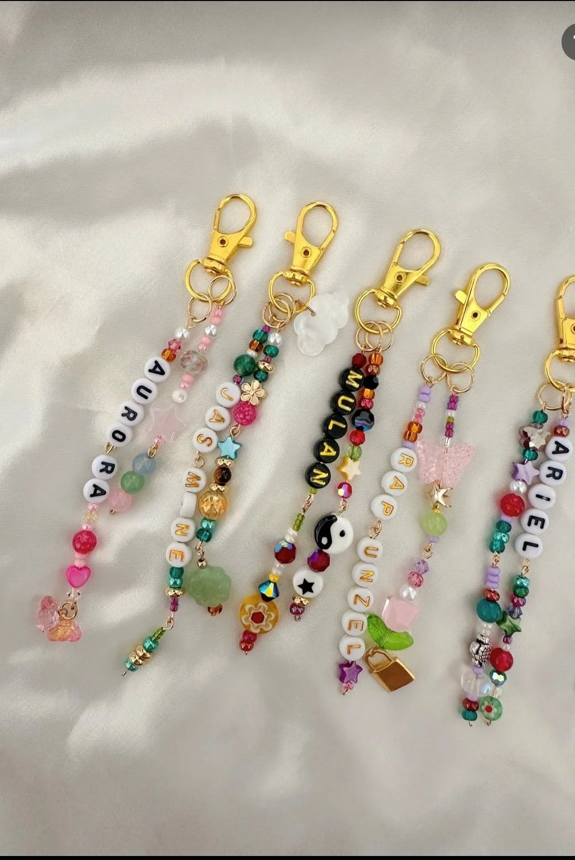 Colorful beaded keychains with gold clips, featuring letters spelling names and various decorative charms, laid out on satin fabric.
