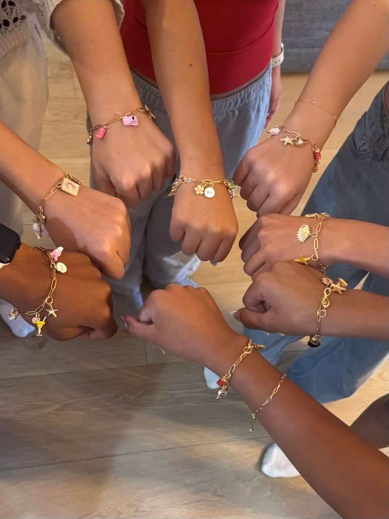 Six people showing their wrists with matching gold charm bracelets, each bracelet has various charms such as a camera, starfish, shell, and other small pendants.