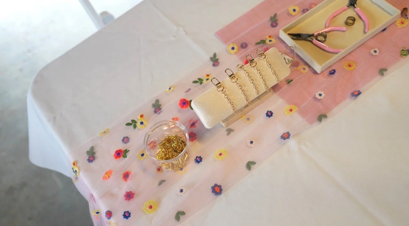 Jewelry display on a table with colorful flower-embroidered cloth, including gold chains, scissors, pink pliers, and rings.