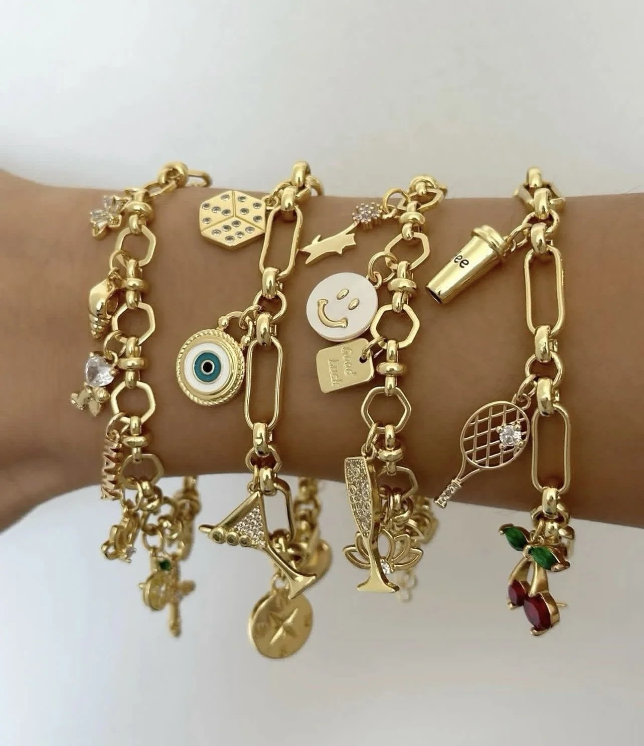 Four gold charm bracelets on a person's wrist featuring various charms including a smiley face, tennis racket, eye, star, and other symbols.