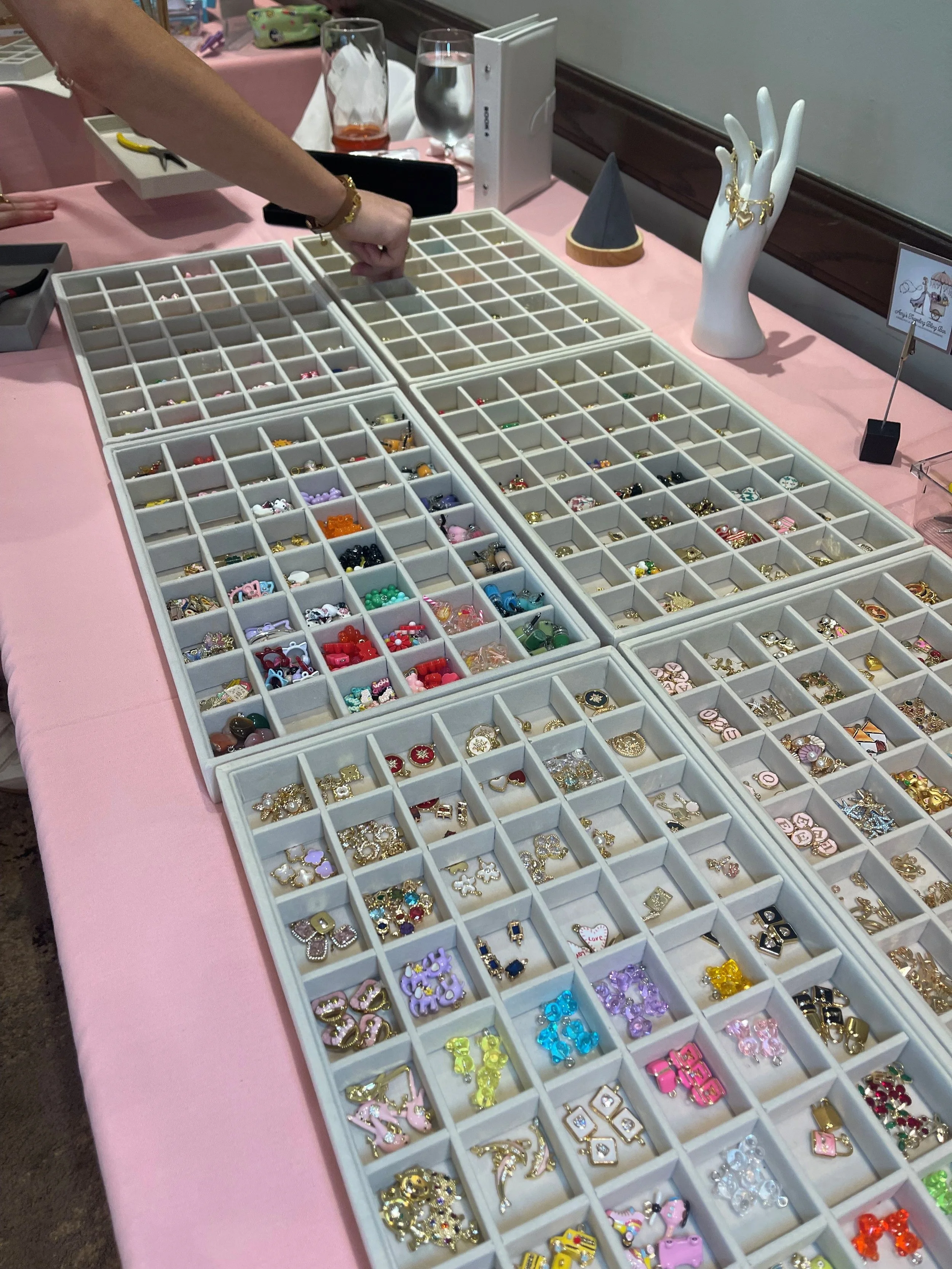 A table displays many small compartments filled with colorful jewelry charms and decorative pieces, with a hand reaching to pick one. There are also jewelry display mannequin hands and glasses on the table.
