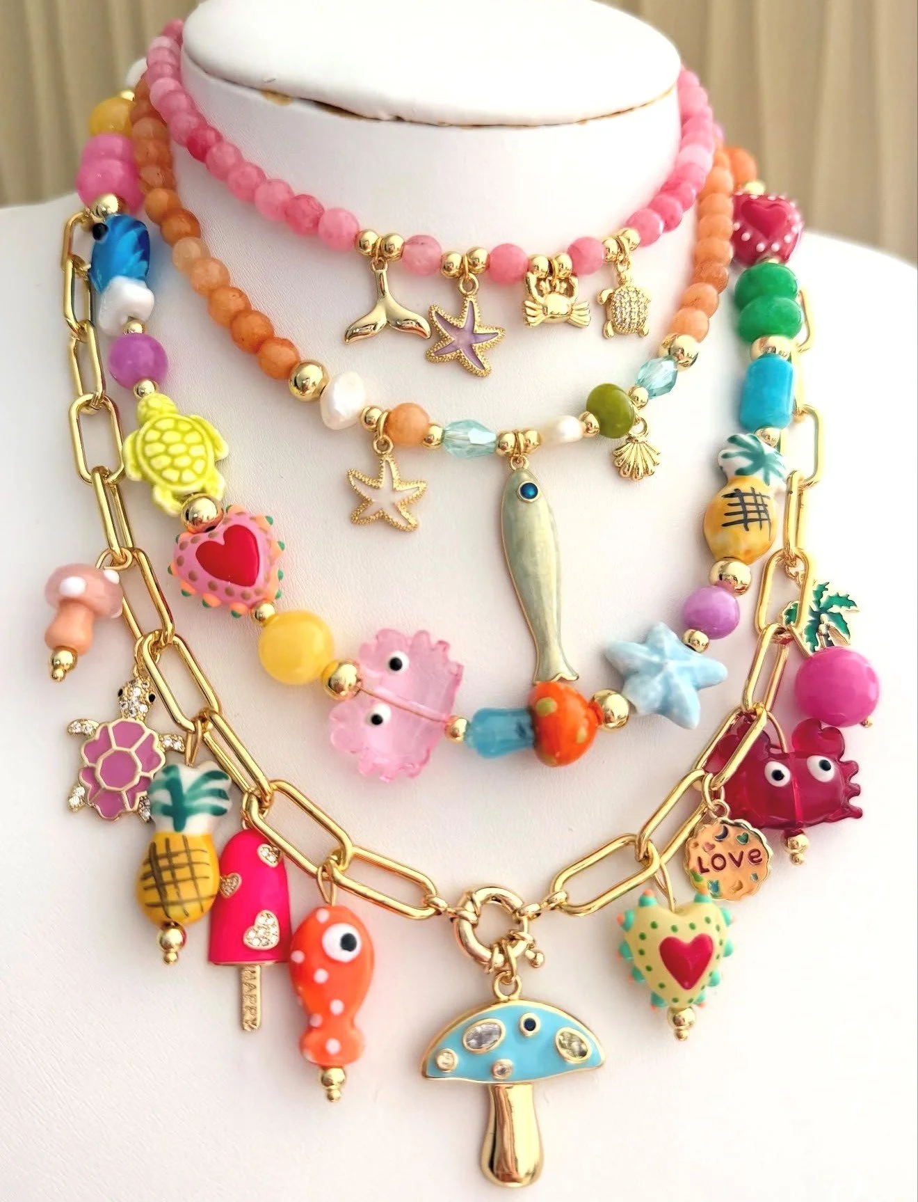 Colorful layered necklaces with charms, including a fish, starfish, seashell, pineapple, mushroom, cloud, heart, and other playful sea and fruit themed designs.