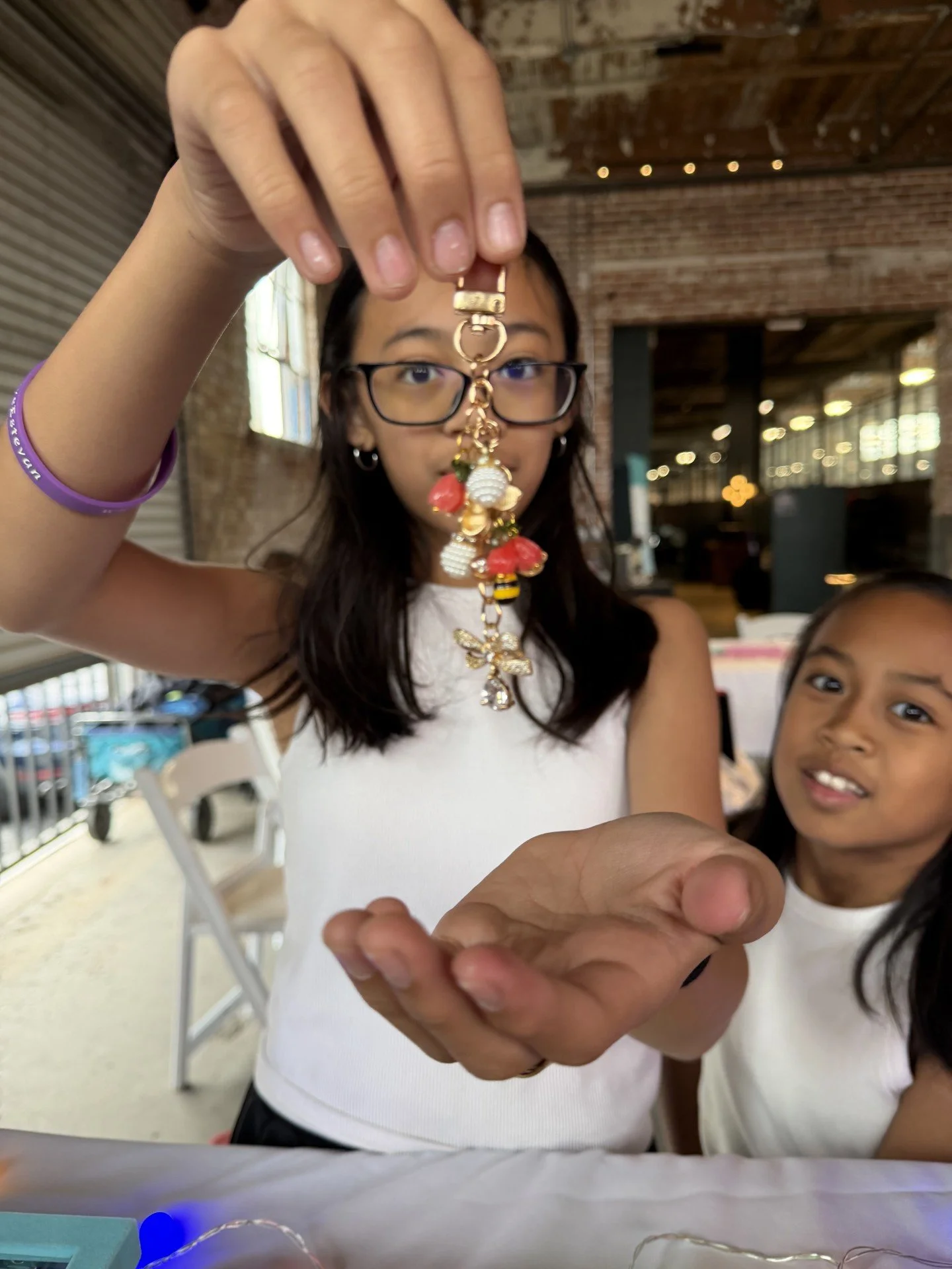 A woman with glasses holding a keychain with various charms, including a bee, a red flower, and a pearl, in front of her face while another girl looks at the camera in a rustic indoor setting with brick walls and string lights.