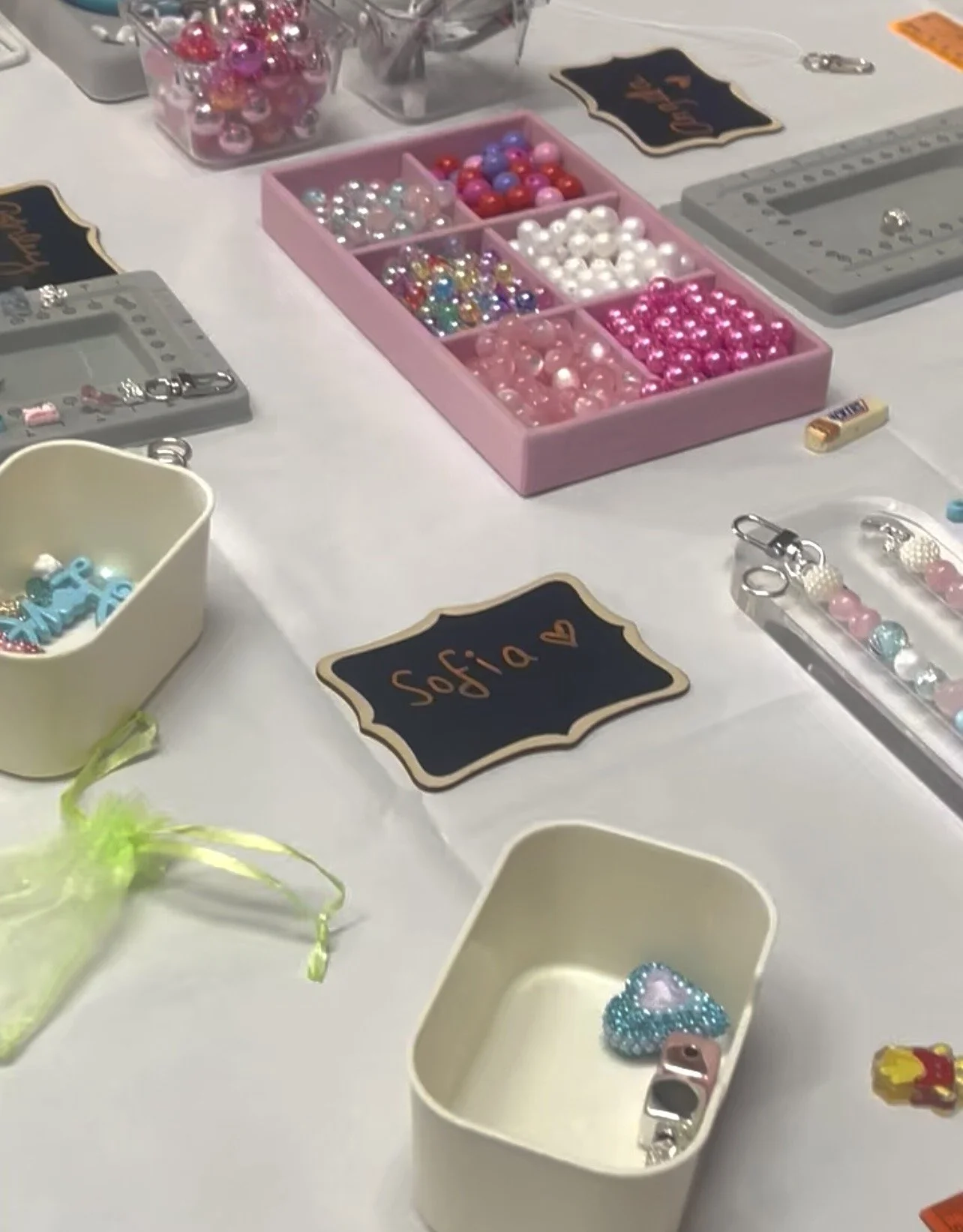 Table displays various colorful beads and jewelry-making supplies, including a pink box with compartments filled with beads, containers with charms and decorations, and a small chalkboard sign with the name 'Sofia' written on it.