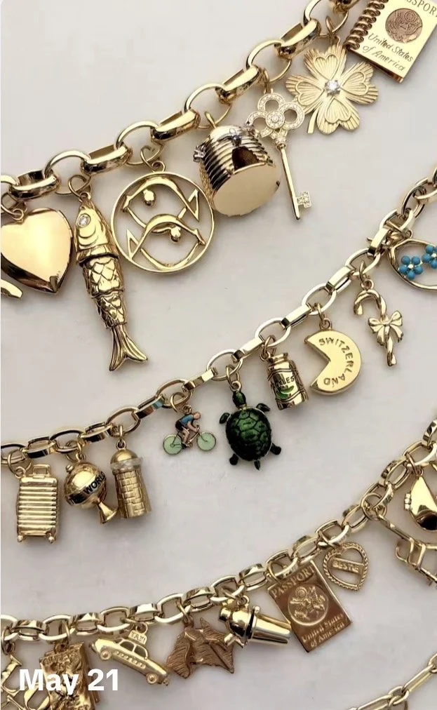 Close-up of four gold charm bracelets with travel, adventure, and miscellaneous charms including a heart, fish, moon and stars, butterfly, turtle, and travel-related charms like a suitcase, passport, and a car.