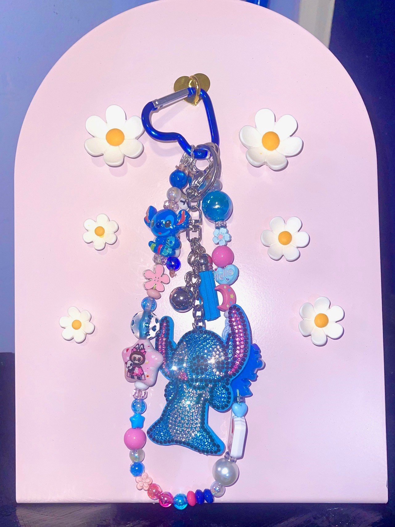 Colorful keychain with a large blue rhinestone dolphin, surrounded by various beads, charms, and a blue clip, hanging against a pink background with five white daisy decorations.