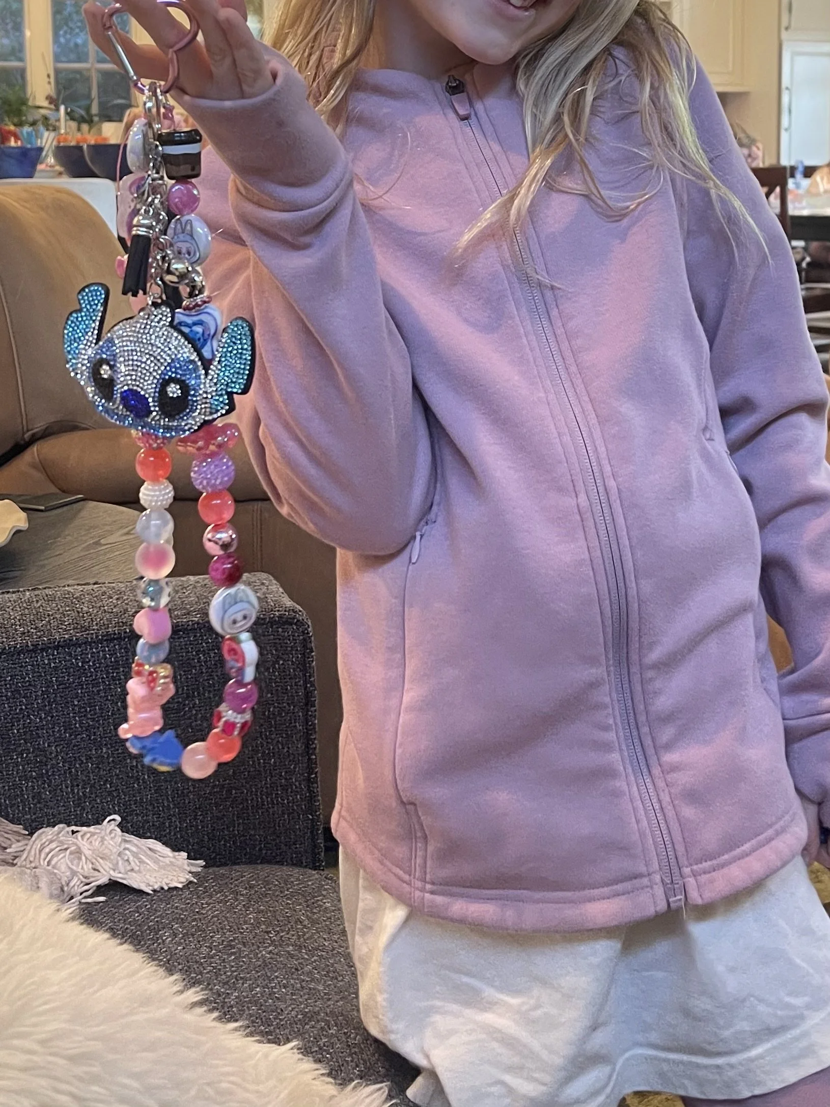 A girl with blonde hair wearing a pink zip-up hoodie and white pants is holding a colorful beaded keychain featuring a rhinestone-studded panda bear. The background shows a kitchen and a living room with a sofa and various household items.