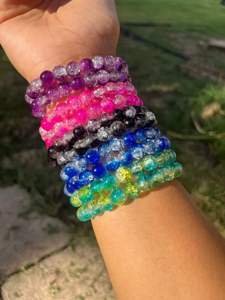 Multiple colorful glittery beaded bracelets worn on a person's wrist, outdoors with grass in the background.