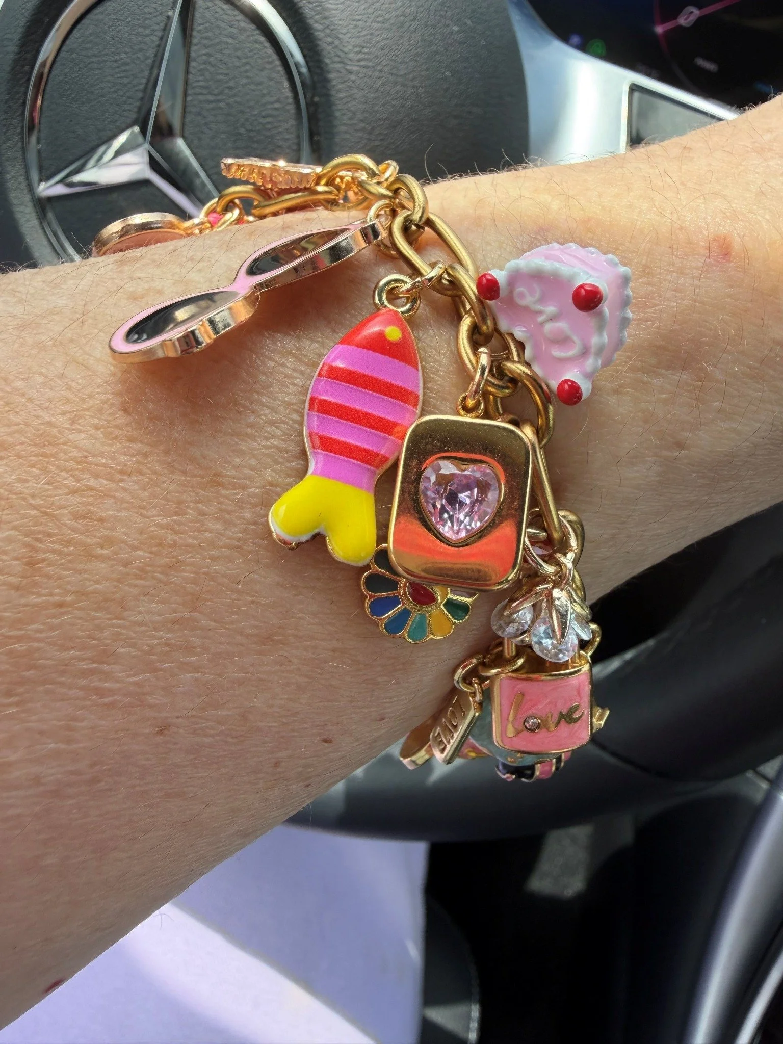 A wrist adorned with multiple colorful charm bracelets resting on a car dashboard with a Mercedes-Benz logo visible in the background.
