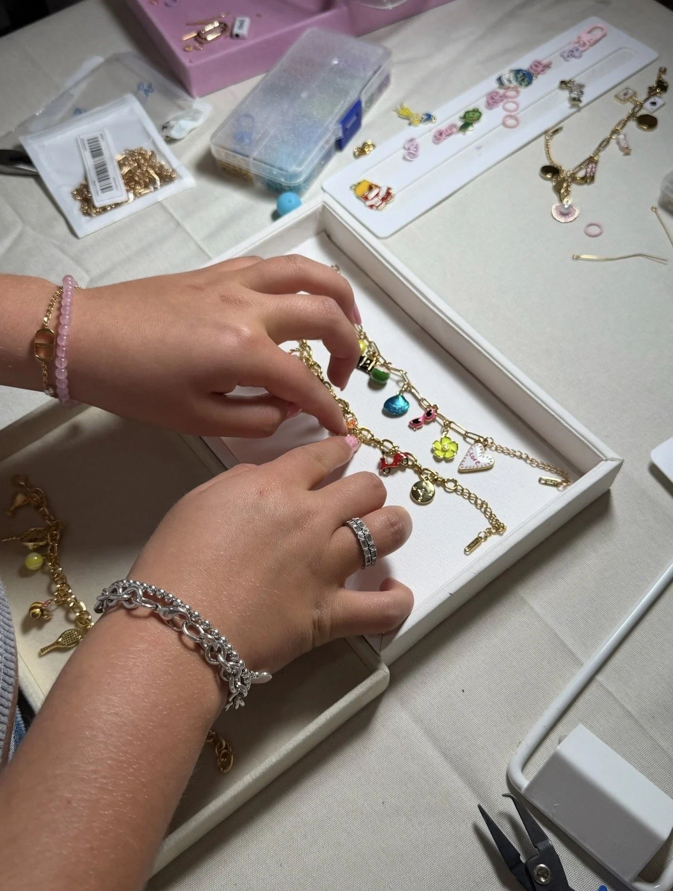 A person handling gold jewelry with various charms and pendants, displaying bracelets and rings, on a white jewelry display box. The workspace includes additional jewelry, a plastic container, and jewelry tools.