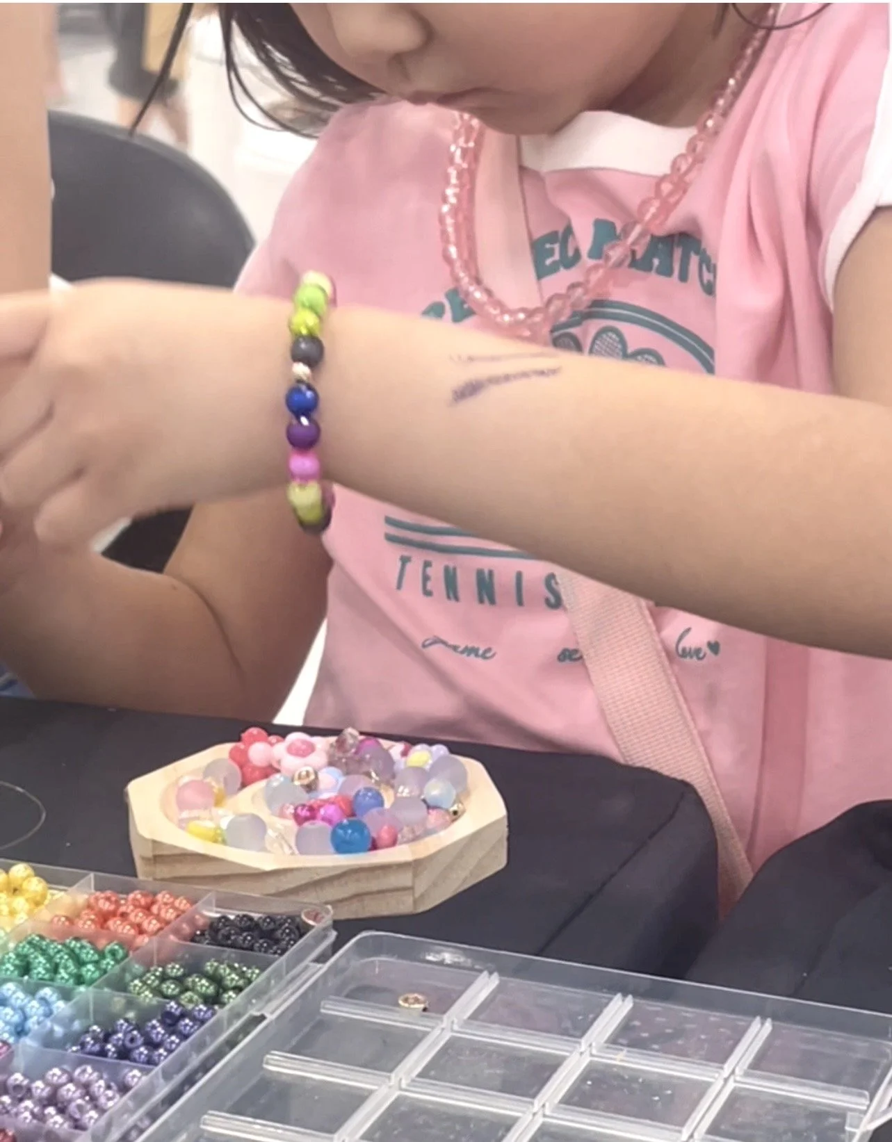 A young girl wearing a pink shirt, pink necklace, and a colorful bracelet, sitting at a table with an assortment of beads and jewelry-making supplies.