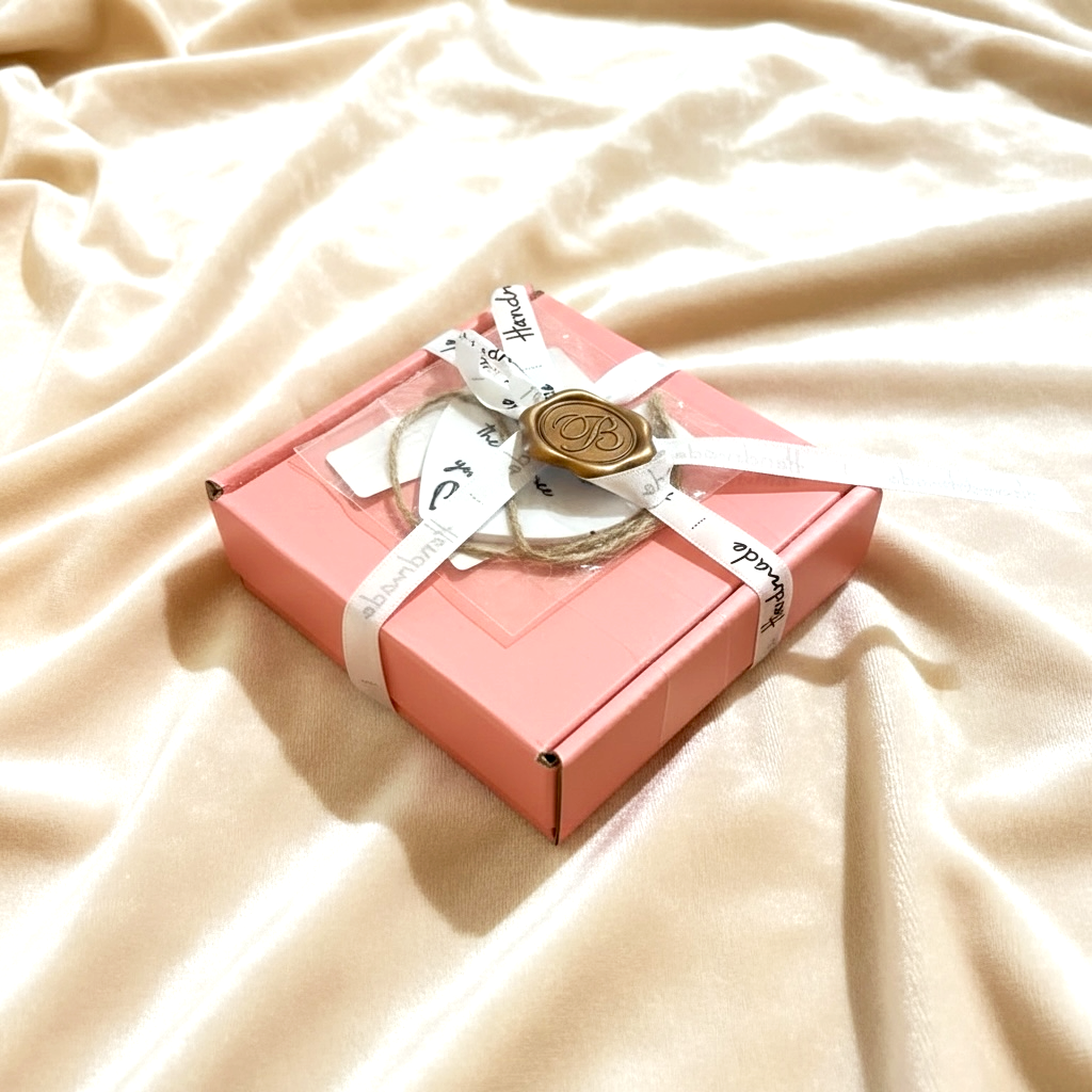 Pink gift box with a white ribbon, a gold wax seal, and tags on a cream-colored fabric background.