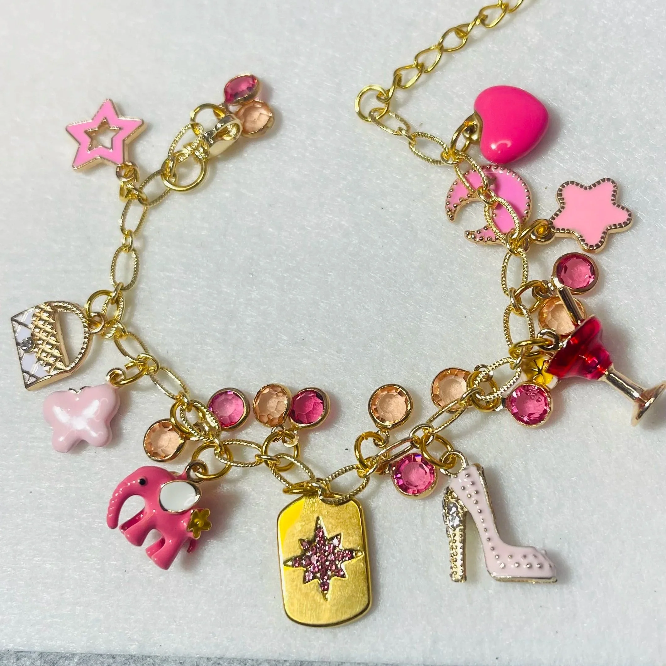 Gold charm bracelet with pink and peach rhinestone accents and charms including a pink butterfly, pink crown, pink star, pink heels, pink elephant, gold tag with purple star, pink heart, and pink starfish.