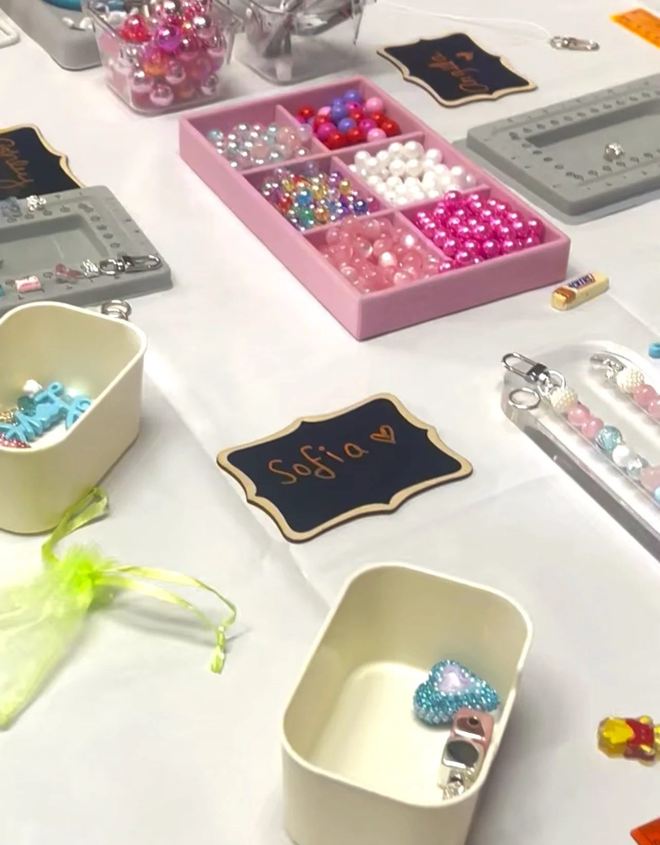 A table with various beads, charms, and adornments for jewelry crafting. There are containers of different colored beads, charms, and a blackboard sign with the name 'Sofia' written on it, indicating a craft or jewelry-making activity.