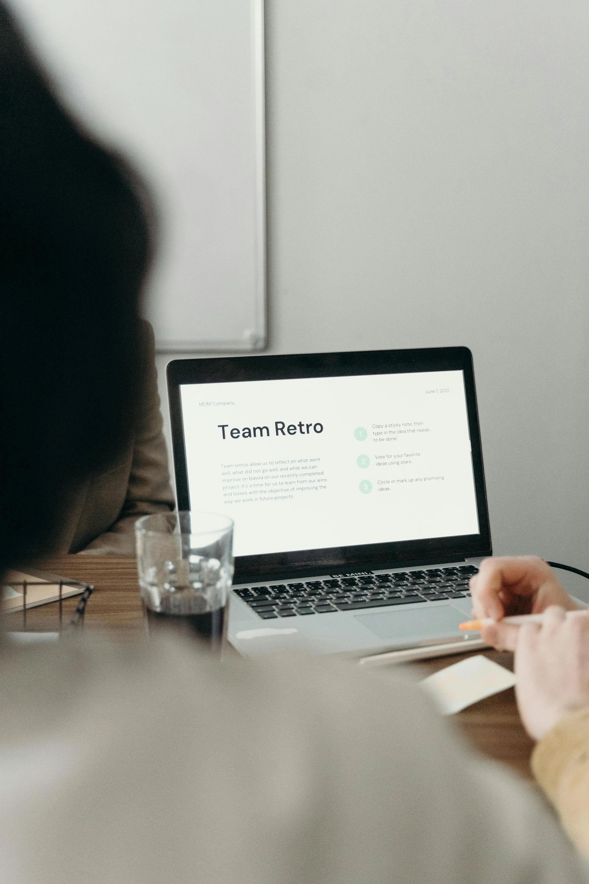 Person working on a laptop with a presentation slide titled 'Team Retro' on the screen, a glass of water nearby, in a meeting or office setting.
