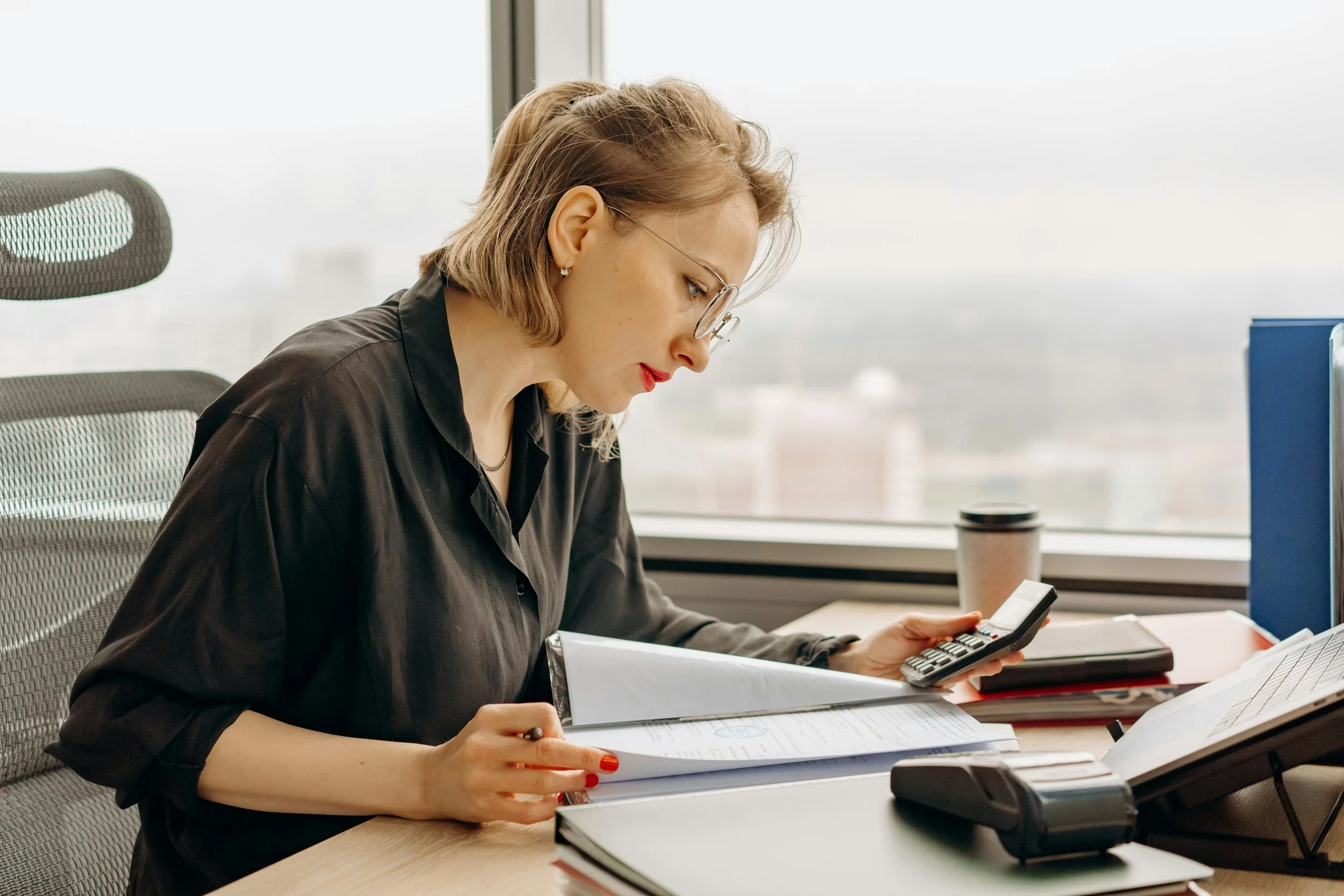 Woman in black blouse working at desk with papers, calculator, and coffee cup.