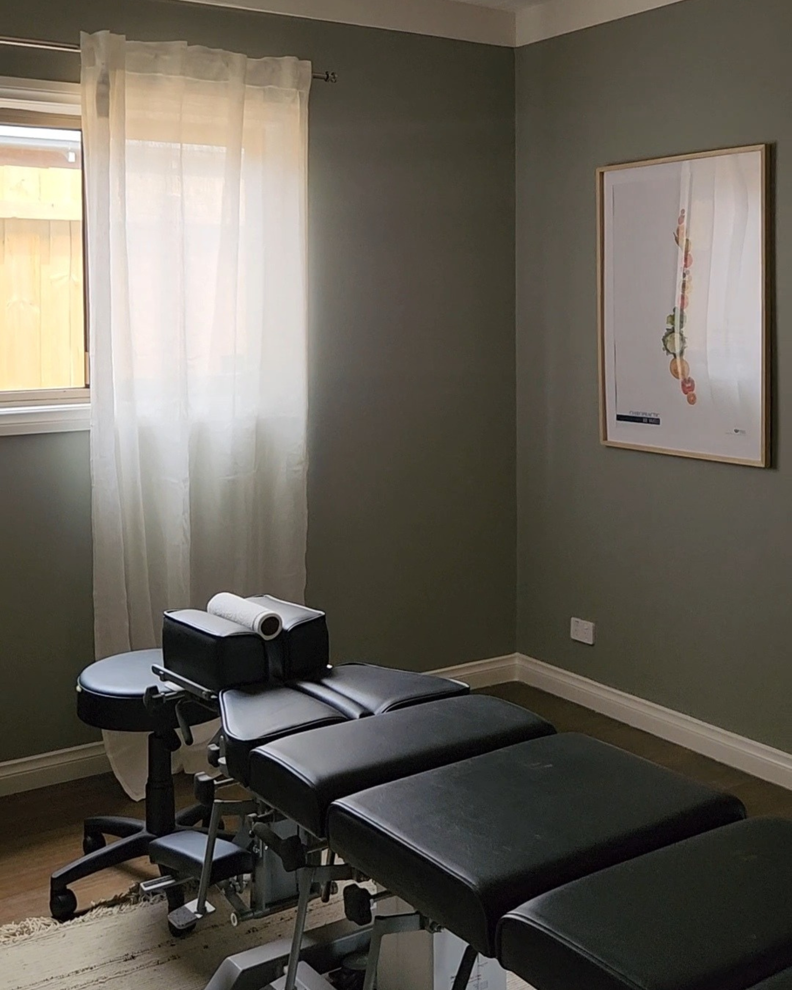 A room with a massage or chiropractic table near a window with white curtains, a framed botanical art print on the wall, and a small rolling stool.