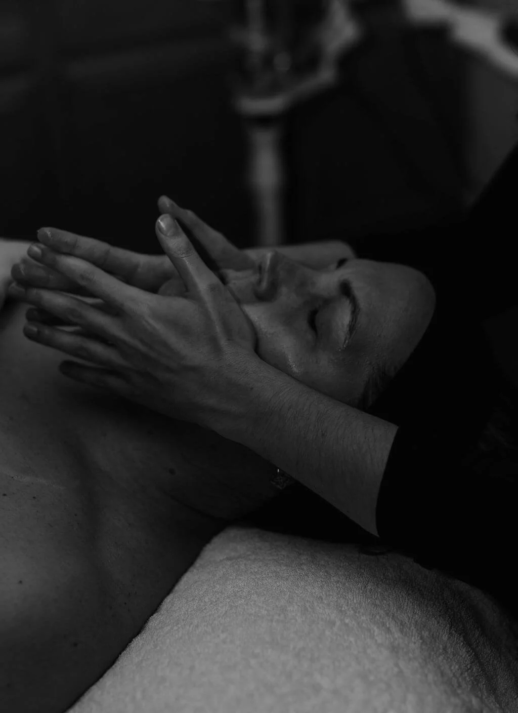 A person receiving a facial massage or treatment, with eyes closed, lying on a surface, in black and white.