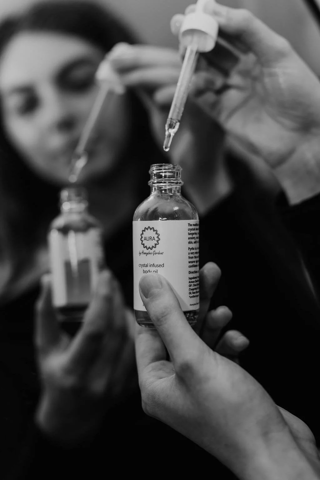 A woman with a face mask and glasses holding a small bottle of crystal infused body oil with a dropper, with another similar bottle in the background.