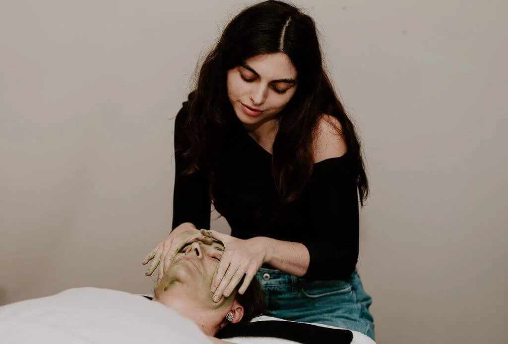 A woman with long brown hair, wearing a black off-shoulder top, performs a facial massage on an older woman lying on a bed or massage table, with her eyes closed, in a calm, relaxed setting.