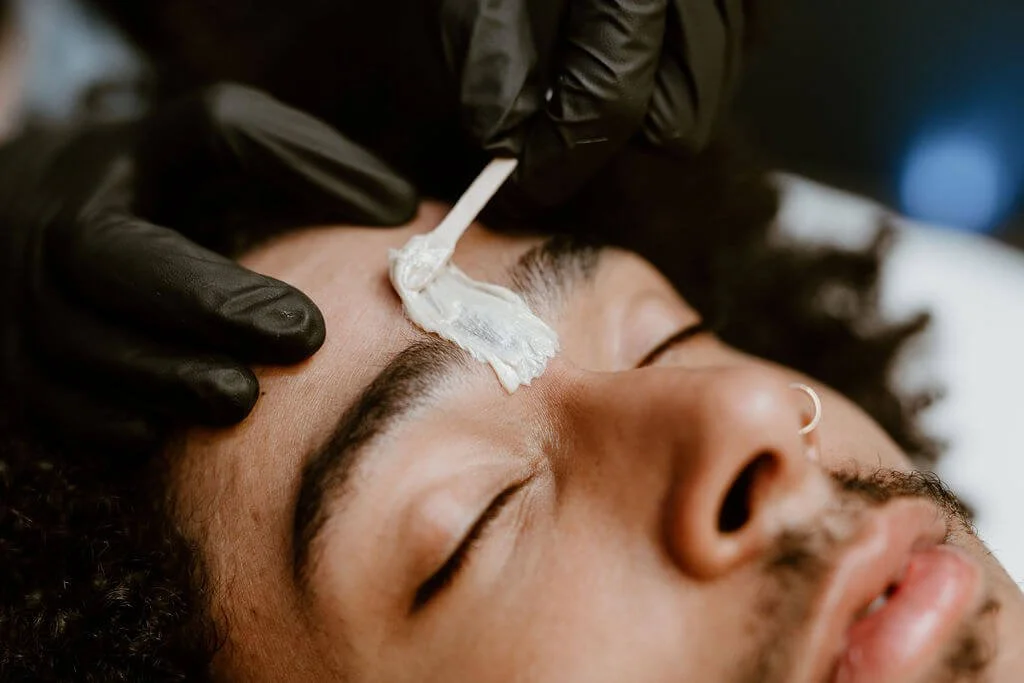 A person lying down with their eyes closed while a gloved hand applies ointment or cream to their forehead with a cotton swab.