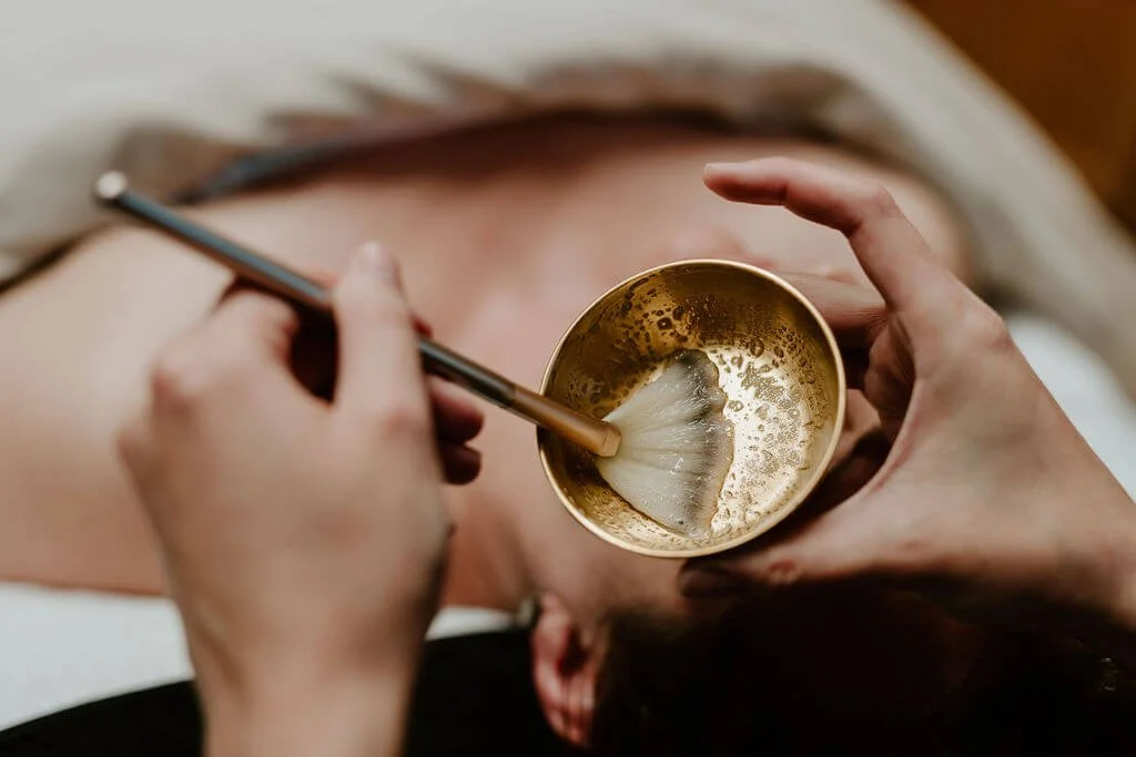 A person receiving a gold facial treatment with a gold bowl and brush