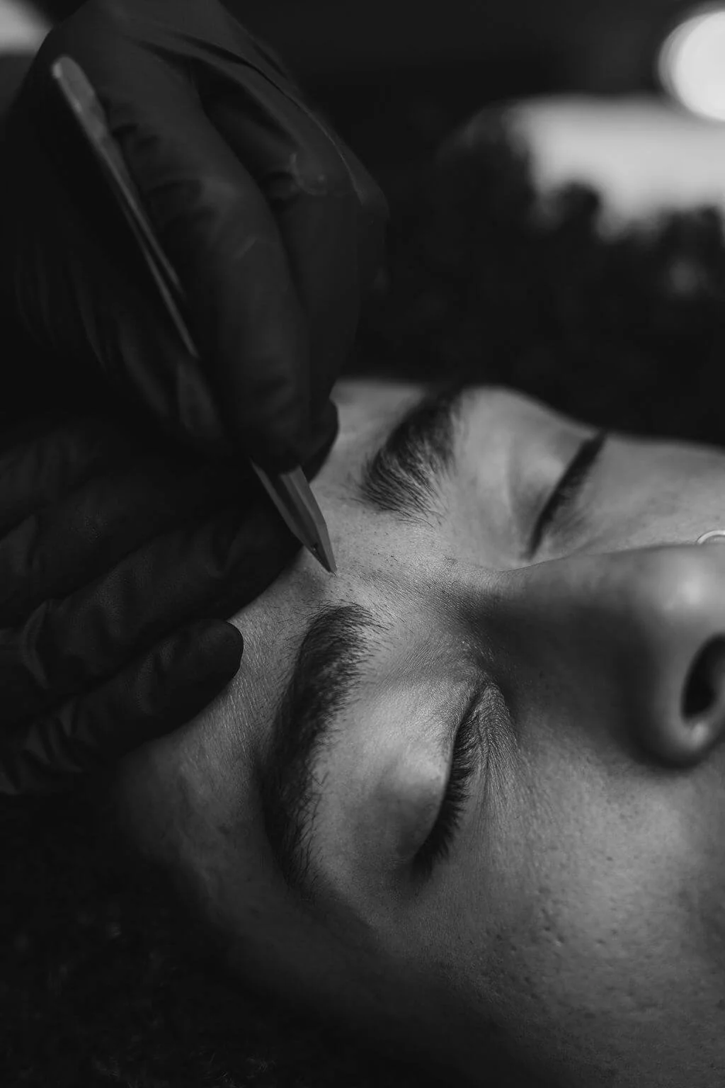 Close-up of a person receiving an eyebrow tattooing or microblading procedure, with a technician wearing black gloves applying ink to the person's eyebrows.
