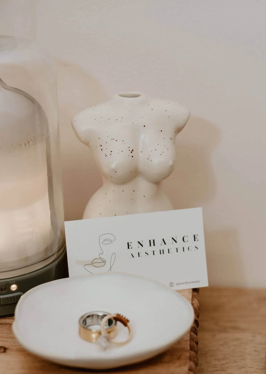 Decorative ceramic torso sculpture, two rings on a plate, skincare or aesthetic branding card, and a white candle in a glass holder on a wooden surface.