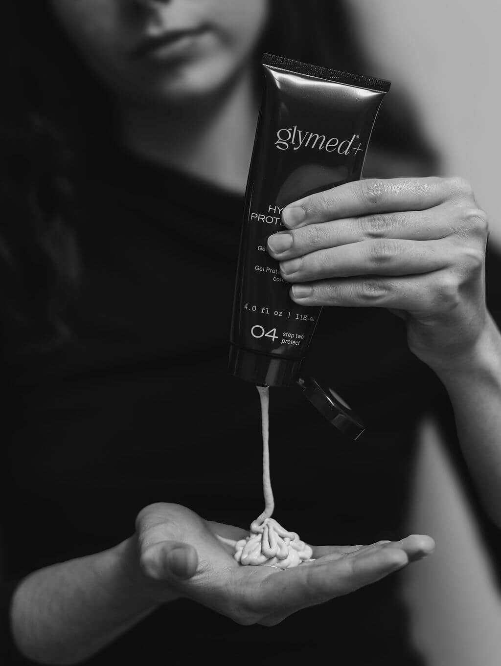 A person holding a tube of Glymed+ skincare product above a palm with a swirl of cream, with cream dangling from the tube.