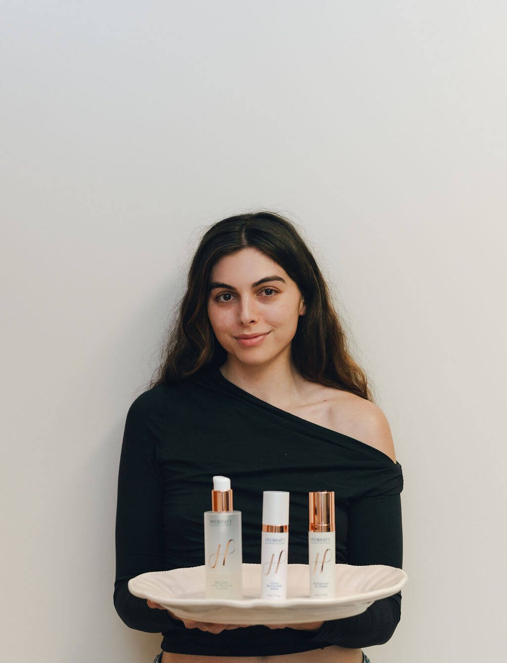 A young woman with dark hair holding a tray with three skincare products.
