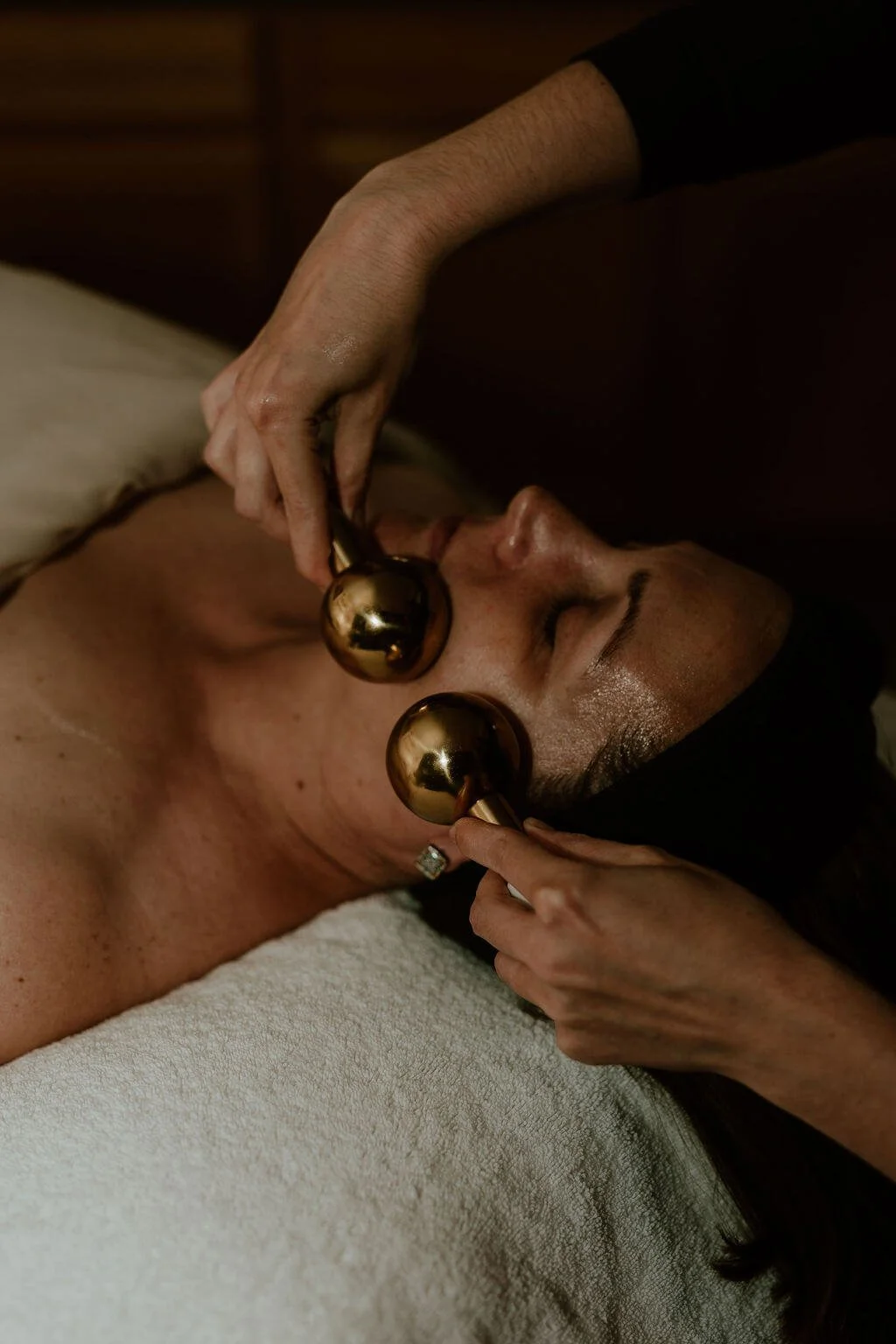 A woman receiving a facial massage with gold facial rollers in a spa setting.
