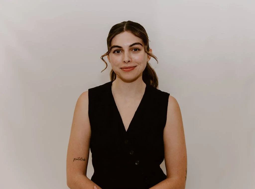 A young woman with brown hair styled in loose waves, wearing a sleeveless black top, standing against a plain light wall, smiling softly at the camera.