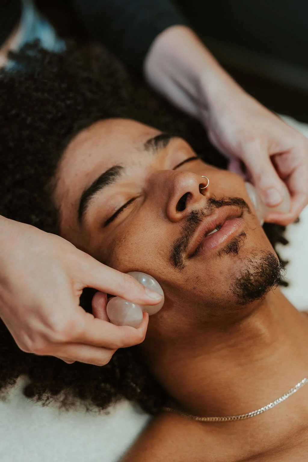 A man with a nose ring relaxing with crystal massage stones on his face at a spa or massage therapy session.