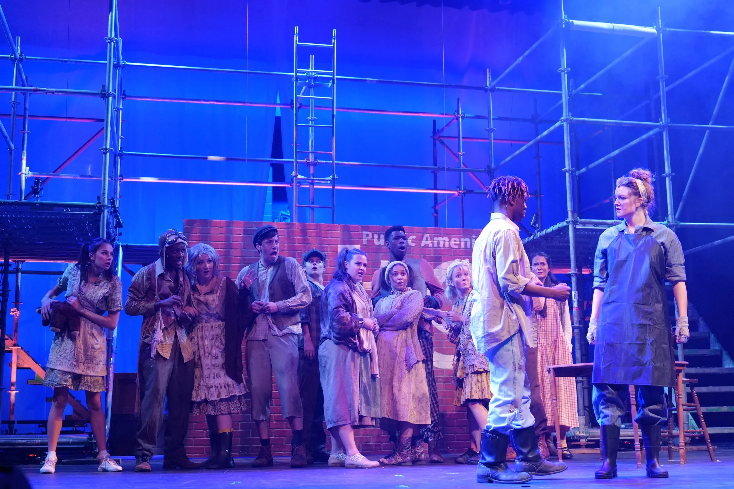 A theatrical stage scene with a group of actors dressed in period costumes, some looking surprised and others engaged in conversation. The set includes scaffolding and a brick wall backdrop with a sign that reads "Public Ameni".