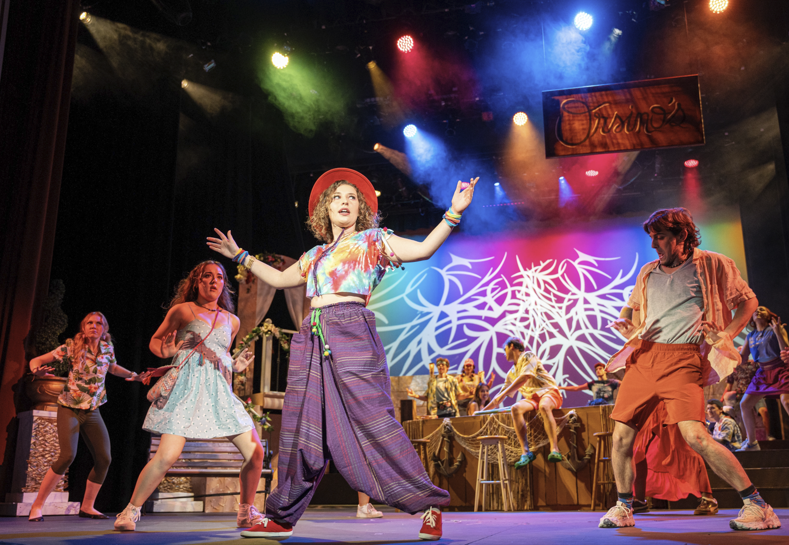 A stage theatrical performance with young actors dressed in colorful casual costumes. The scene is lively with a woman in the center wearing a tie-dye crop top, wide pants, and a red hat, raising her arms. The background features vibrant lighting, and a sign that reads 'Ovise' is hanging above. The set includes props like chairs and barrels, with a decorative backdrop of white abstract designs on a rainbow gradient.