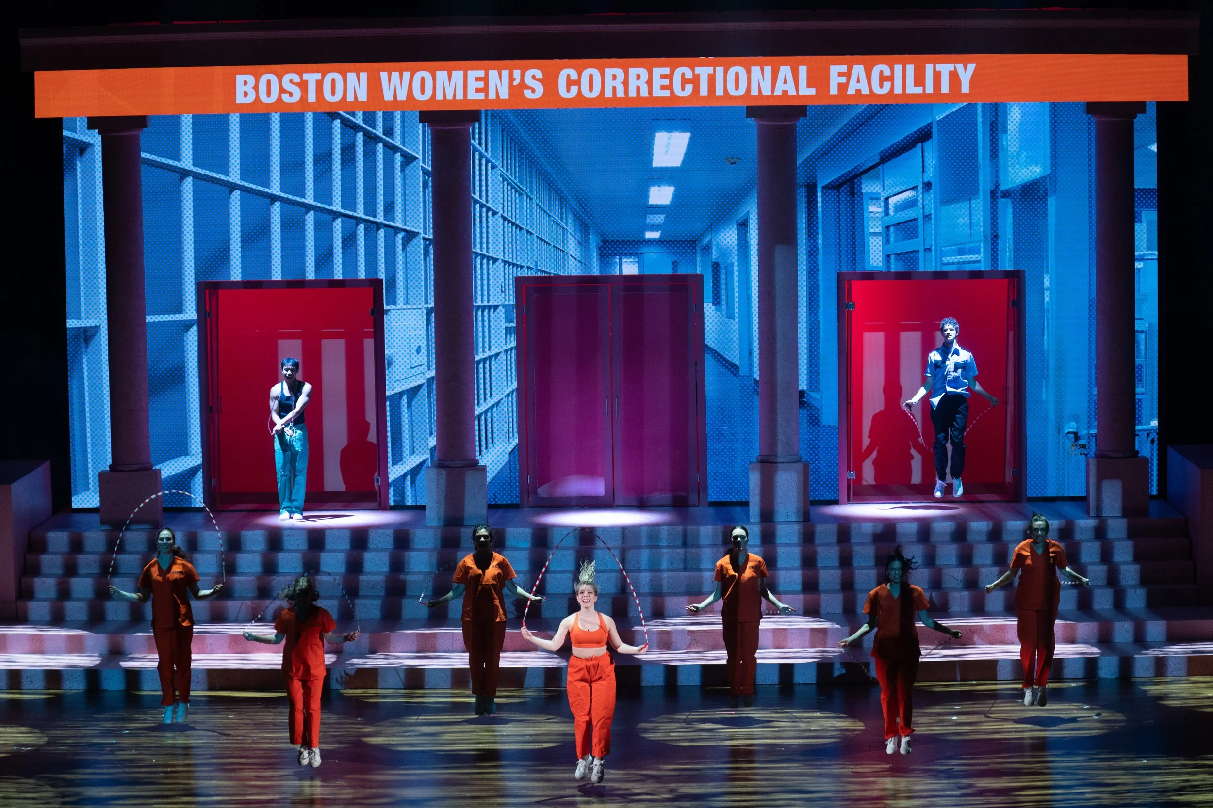 A stage performance with a large digital backdrop displaying the text 'Boston Women's Correctional Facility.' Dancers dressed in orange scrubs and masks perform a coordinated jump, while two others appear to be emerging from red doors on the set.