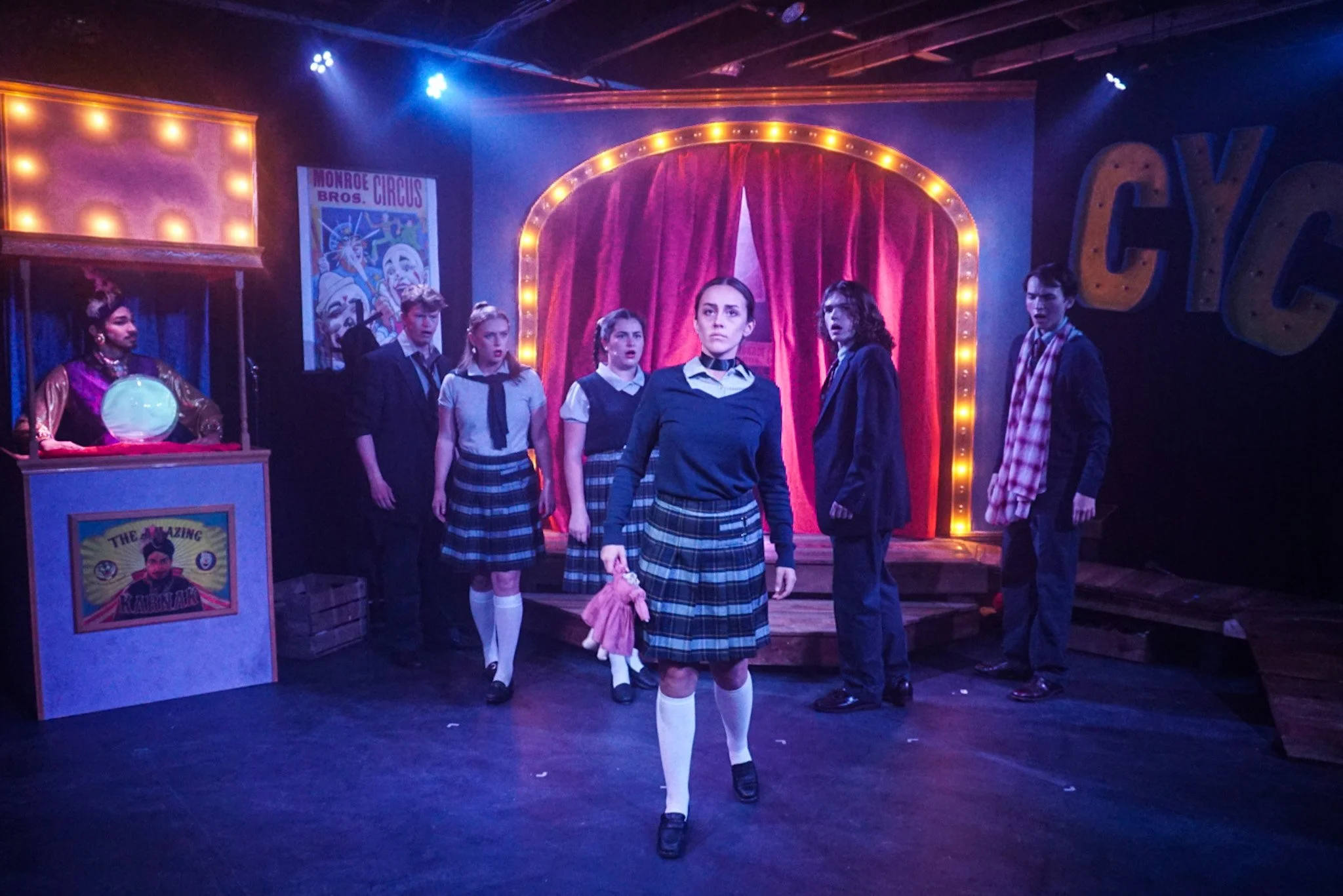 Students in school uniforms on stage during a performance at a carnival-themed theater with a red velvet curtain, illuminated letters reading 'CIRCUS', and a poster for Monroe Bros. Circus.