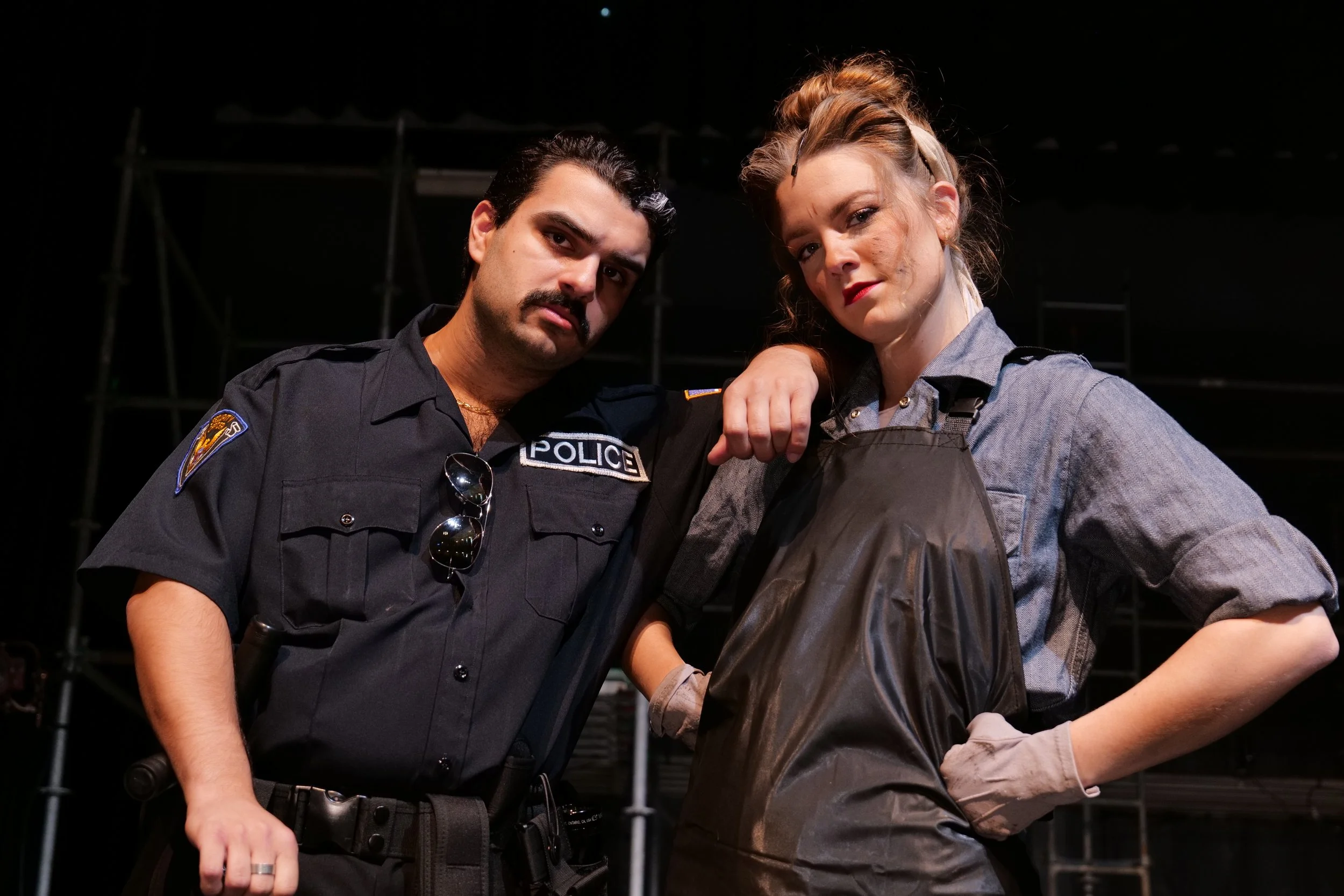 Two people, a man dressed as a police officer and a woman in a gray shirt with an apron, standing close together with their arms on each other's shoulders, looking serious. They are indoors with a dark background.
