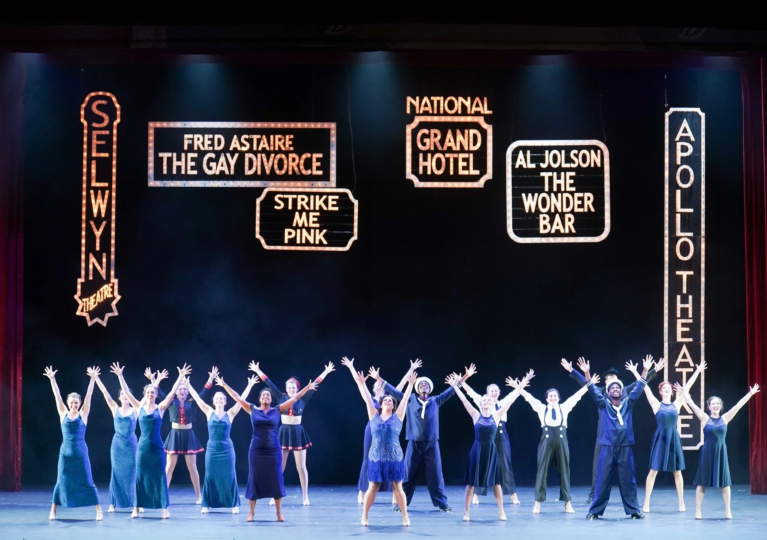 A theatrical stage with cast members taking a bow, dressed in vintage costumes, under illuminated signs displaying theater and show titles such as 'Hello, Dolly!', 'Fred Astaire The Gay Divorce', and 'Grand Hotel'.