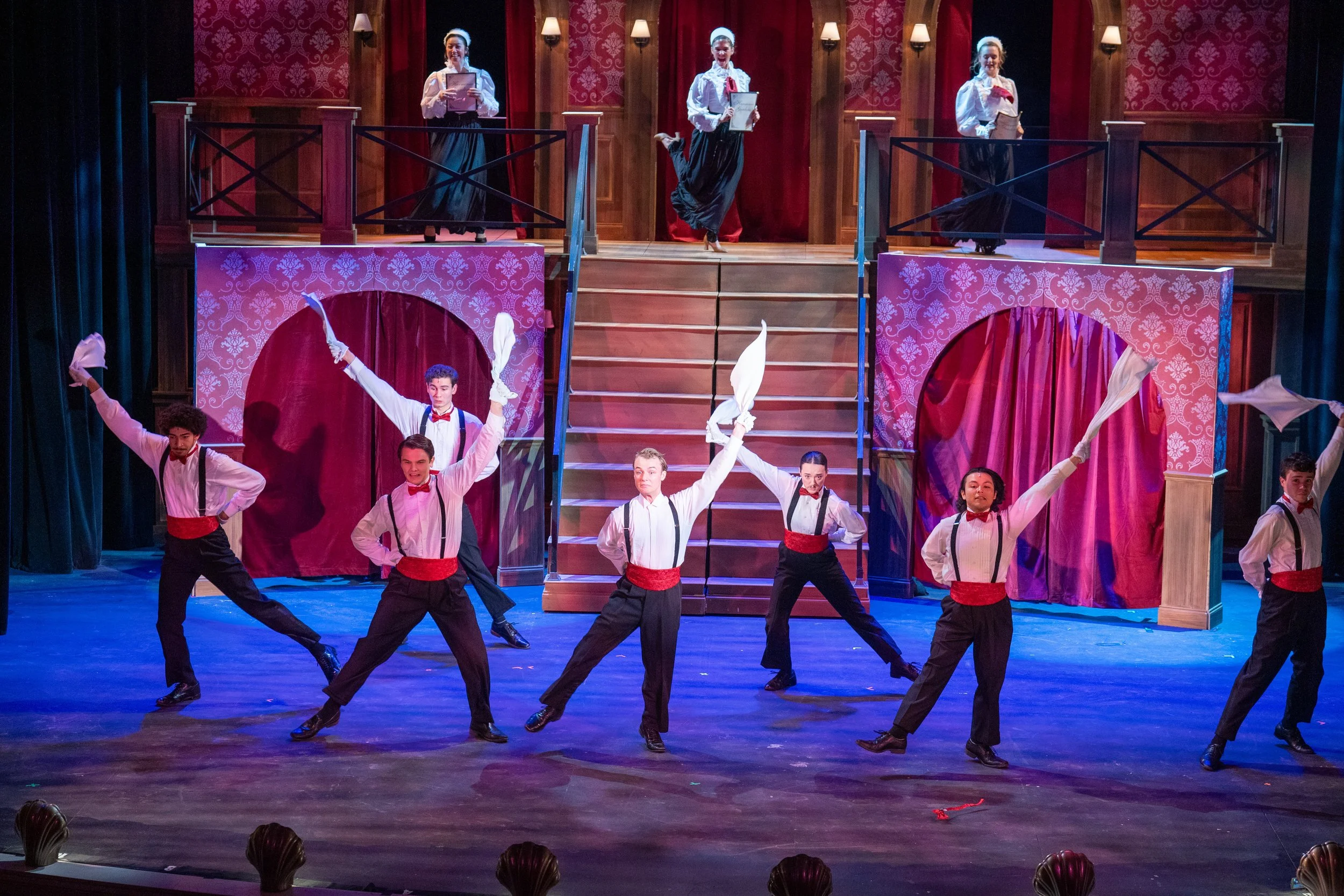 The stage scene from a musical with a group of dancers in black pants, white shirts with suspenders, and red sashes, performing with white handkerchiefs, while four women in historical costumes stand on a balcony above.