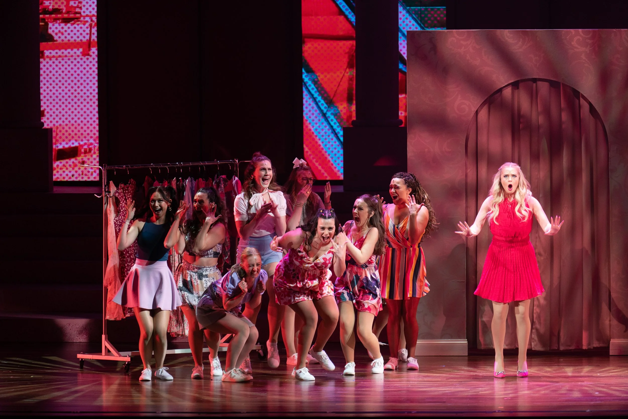 A group of women performing on stage dressed in colorful summer outfits, appearing excited and expressive, with some holding hands and others with hands near their faces, in a theatrical production setting.