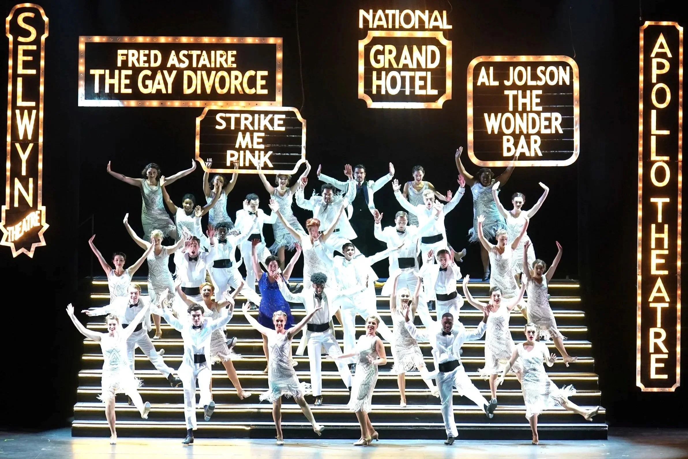 A theatrical stage with a large cast of performers in white costumes, dancing on steps with colorful signs in the background displaying titles and names of shows, including 'Fred Astaire The Gay Divorce,' 'Strike Me Pink,' 'Al Jolson The Wonder Bar,' and others, illuminated with bright lights.