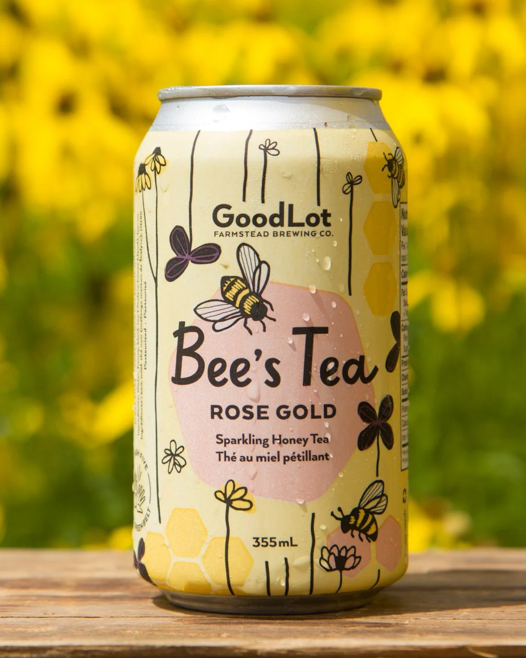 A can of Good Lot Farmstead Brewing Co.'s Bee's Tea Rose Gold sparkling honey tea with illustrations of bees, flowers, and honeycomb on the label, sitting on a wooden surface outdoors with a yellow floral background.