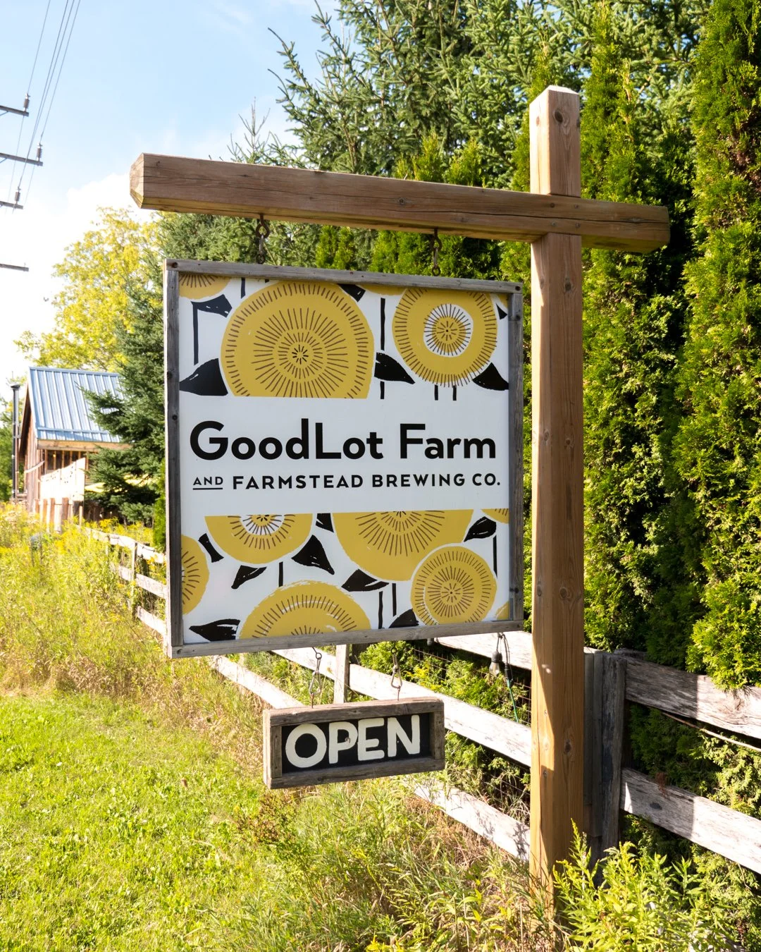 A sign for GoodLote Farm and Farmstead Brewing Co., with a yellow sunflower design, and an 'OPEN' sign hanging below, outdoors with green trees and a wooden fence.