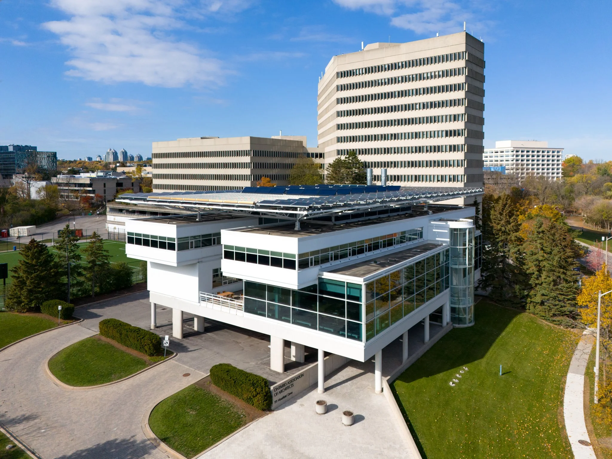 Modern white building with large glass windows and solar panels on the roof, surrounded by greenery and trees, with taller office buildings in the background under a clear blue sky.